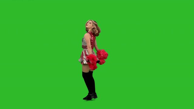 An adorable cheerleader in uniform with red pom-poms performs a jubilant dance. Portrait of a young smiling woman dancing in the studio on a green screen. Slow motion.