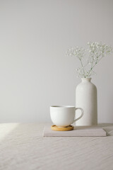 Cup of coffee and vase with gypsophila flowers on table with linen tablecloth. White wall on background. Still life nordic, Scandinavian home decor