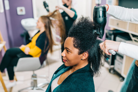 A hairdresser dries a Black woman's hair in a modern hair salon. Beauty, fashion concept. Caucasian client in a background.