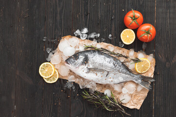 Raw dorada fish or gilt-head bream on ice with lemon slices and rosemary over black wooden background, flat lay, top view.