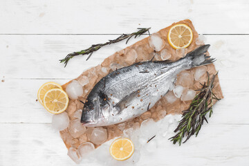 Raw dorada fish or gilt-head bream on ice with lemon slices and rosemary over white wooden background, flat lay, top view