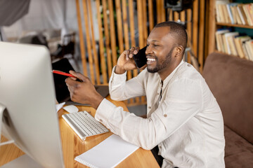 Happy male freelancer sitting at the desk in home office, having a phone conversation with colleague, smiling. Stylish african american businessman discussing ideas for new project or startup