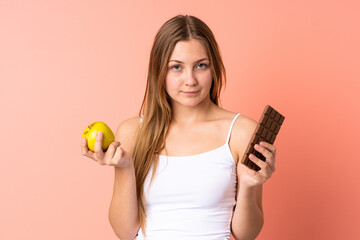 Teenager Ukrainian girl isolated on pink background taking a chocolate tablet in one hand and an apple in the other