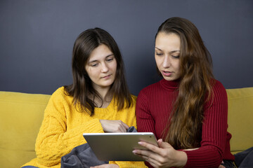 two pretty long-haired brunette girls make purchases or payments online, female millennial sit relax on yellow sofa at home using tablet and internet for Couch shopping on ecommerce site