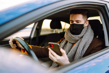 Male driver in protective medical mask used mobile phone sitting in a car. Driver on a city street during a coronavirus outbreak. Covid-19.