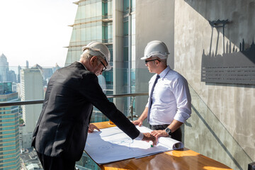 Two engineers discuss their project. They wear white shirt and suit. They also wear white hard hat. They use a pen and paper to share their idea.