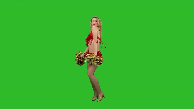 A cheering dance performed by a cheerleader in a red leotard with pompoms in her hands in the studio on a green screen. A charming girl moves her arms and hips, twirls and waves her hair. Slow motion.