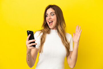 Caucasian woman isolated on yellow background looking at the camera while using the mobile with surprised expression