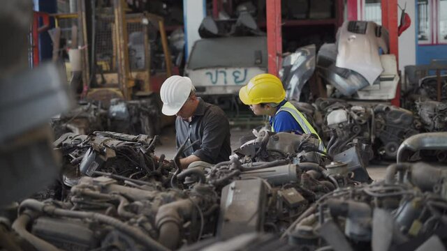 senior engineer using laptop computer checking list old used car engine with young asian worker in machinery parts shop warehouse. old manager find vehicle automobile machine in Recycle motor factory
