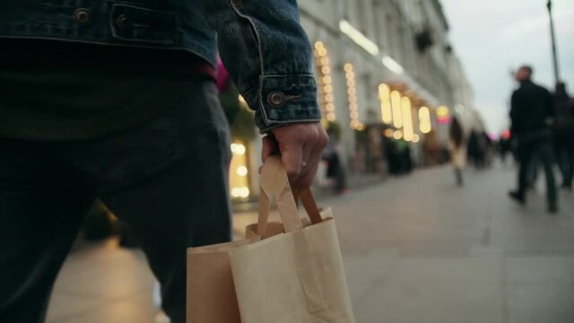 man's hand in a denim jacket carries a yellow recyclable paper eco-friendly bag down a city street. Evening on a crowded street in the center of the metropolis. Shopping alone. Postal delivery 