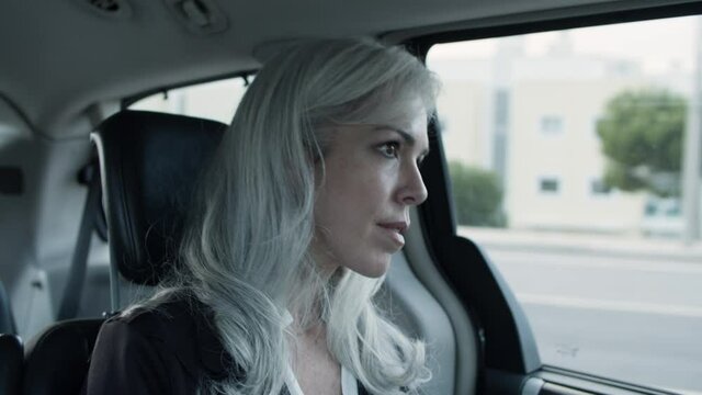 Gray-haired woman in official suit sitting in moving car. Medium shot of middle-aged lady looking out window from auto. Businesswoman, taxi, transport concept.