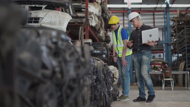 senior engineer using laptop computer checking list old used car engine with young asian worker in machinery parts shop warehouse. old manager find vehicle automobile machine in Recycle motor factory