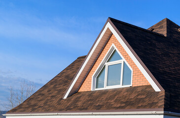 Roof of house is with a triangular dormer window. Flexible roofing material in brown color. Close-up at an angle. Background - blue sky. Concept of a new cottage.