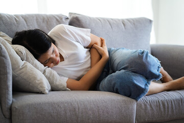 Young asian woman suffering from strong abdominal pain stomachache or period comes while lying on sofa at home