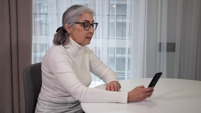 Happy old senior grey-haired woman grandmother waving hand talking with daughter on smartphone video conference calling enjoying social distance party, virtual family online chat meeting at home.