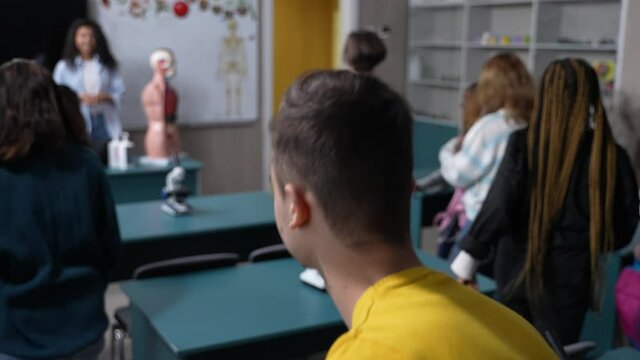 Multinational secondary school students getting up from desks and leaving classroom at the end of biology lesson. Mixed race female teacher saying goodbye to group of diverse teen pupils after classes