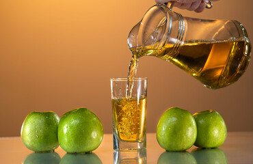 Fresh apple juice is poured from a carafe into a glass. Nearby are green apples on the reflective...