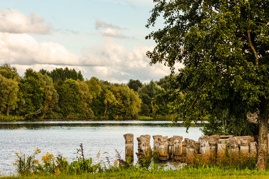Stone bridge pillars ruins in Daugavas river water during sunny summer day framed by green and yellow grass and green trees. Scenic photo of pier remains in water. Sunny day at riverside. © Evelyn