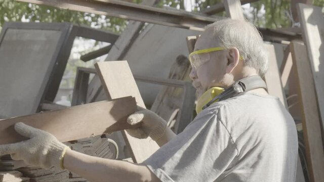 Old man carpenter carefully looking and inspecting wood plank in the factory.