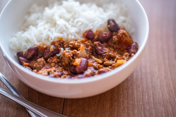 Close up and selective focus of meat chilli concarne and white rice in a white bowl on a wooden surface with intentional shallow depth of field and bokeh
