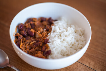 Close up and selective focus of meat chilli concarne and white rice in a white bowl on a wooden surface with intentional shallow depth of field and bokeh