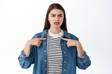 Offended and upset girl points at herself, frowns and looks hurt or insulted, who me gesture, being...
