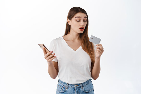 Image of girl looks curious at bank credit card, holds smartphone in hand, pays for online order, shopping in internet store, standing over white background