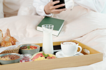 Cup of coffee, glass of non-dairy yogurt, fruits, bread and granola bowl on wooden tray in front of...