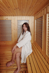 Pretty young Asian woman in bathrobe sitting on bench in hot sauna