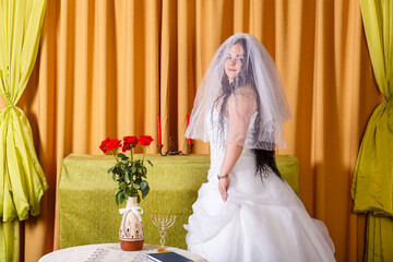 A happy Jewish bride in a fluffy white dress, her face covered with a veil, stands in the room at the table before the chupa ceremony.