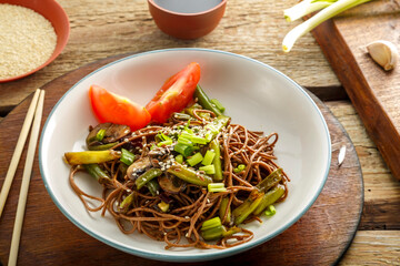 Soba with mushrooms, green beans and sesame seeds in a plate on a stand on a wooden table next to chopsticks and soy sauce and garlic on stands.