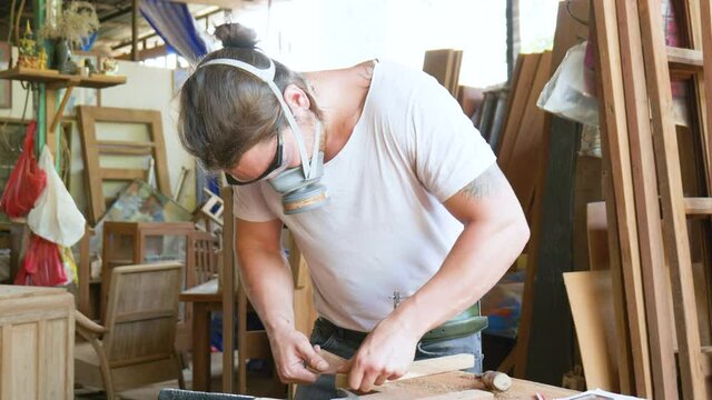Carpenter wearing mask and working on wood plank in workshop of factory.