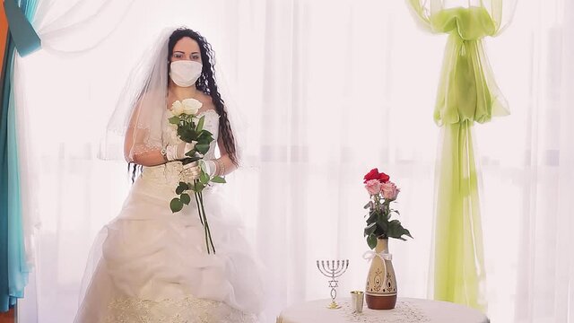 A Jewish bride in a wedding dress and a veil in a medical mask in the synagogue hall with a bouquet of white roses turns her face.