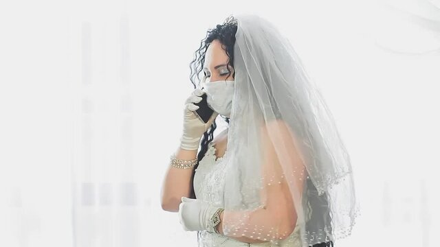 A Jewish bride in a wedding dress and veil wearing a medical mask in a synagogue hall before the chupah ceremony speaks on the phone