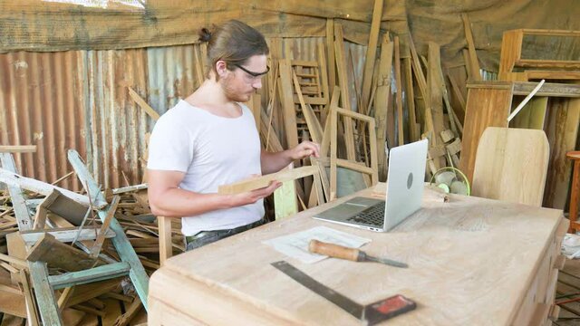 Carpenter working on laptop and holding a wood plank in factory.