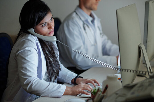 Serious young female Indian doctor talking on phone with patient and entering his complaints and symptoms in form on computer scren