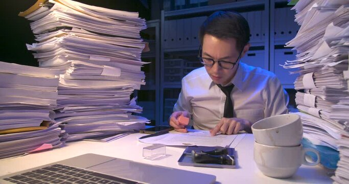 Busy asian business man working multi tasking, talking phone, using laptop, doing paperwork. Tired alone male have a headache while working hard overload at late night.Time lapse, Fast speed shot.