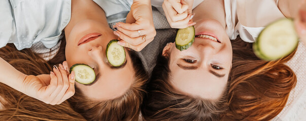 Two girls make homemade face and hair beauty masks. Cucumbers for the freshness of the skin around the eyes. Women take care of youthful skin. Girlfriends laugh at home lying on the floor on pillows.