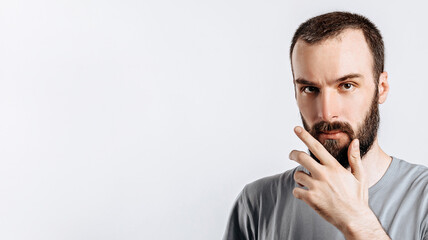 Serious puzzled handsome male model with beard holding hand on chin as if thinking about something, squinting at camera with suspicious look and standing over gray background. Man decides what to buy