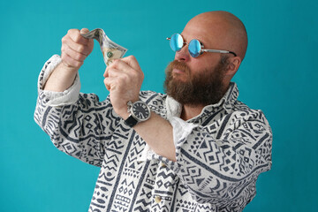 A guy with a bald head and a lens in his hands examines the money. A man with a full beard and mustache in a colorful shirt. Man portrait with glasses.                               