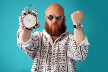 A man with glasses and a beard with a wrist watch holds an alarm clock in his hands. A bald-headed...