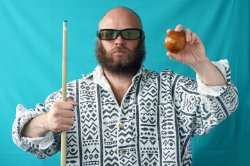 A man with glasses holds a brown billiard ball in his hand. A guy with a bald head and a lush beard in a shirt holds a cue. Sports game. Billiard player                              