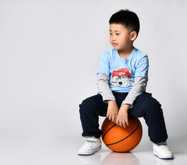 portrait of a preschool little smiling asian boy wearing a trendy raglan with a cool wolf print, sitting on a big orange basketball ball and waiting to be played with