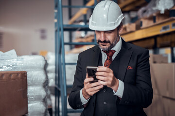 Businessman wearing a hardhat standing at goods warehouse and check for control loading from Cargo freight ship for import and export by report by smartphone. commercial warehouse for trading concept