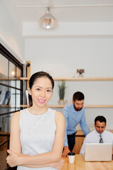 Portrait of positive young businesswoman smiling at camera, her coworker discussing documents on laptop in background