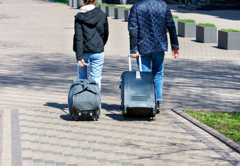 A young couple of travelers walk along the cobbled sidewalk, carrying a travel bag and suitcase on wheels.