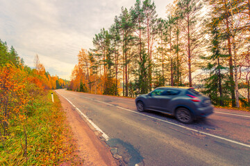 Turn the country broken road. The car goes on the road. Mixed forest. Sunset over the forest lake. Autumn weather. Beautiful nature. Russia, Europe. View from the side of the road.