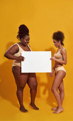 Full length shot of two cheerful african american women in underwear with different body weight smiling, holding blank banner for your text, standing isolated over yellow background