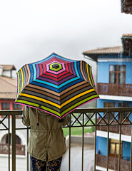 Young woman in khaki jacket from behind with umbrella in rainbow pattern in drops standing on the terrace in the rain, rainy weather in autumn day