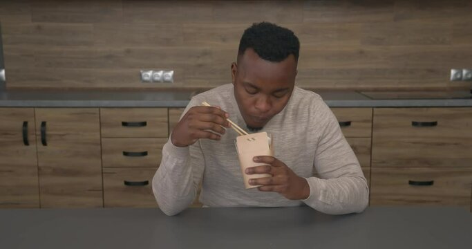 African man sitting at the table and eating with pleasure Chinese noodles from box with chopsticks. Asian street food. Take away